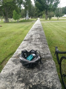 A photo of a black hemet on the wall aroun a cemetary. A black bicycle leans against the wall.