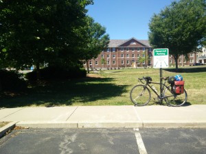 A bike, loaded with gear, leaning against a sign. Behind it is a large, historical hall, and to the left a tree.