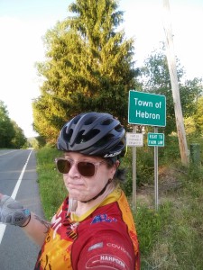 A photo of a cyclist, wearing a red and orange cycling jersey, pink sunglasses, and a black helmet, shrugging in front of a road sign that says "Town of Hebron" and "Right to Farm Law." There is a field, a tree, a telephone pole, and a road in the background.