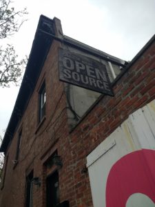 A brick buildig with a wooden sign that says "Come in we're open source!"